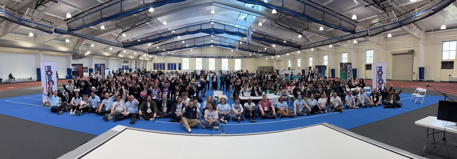 Several hundred competitors, mentors and volunteers sitting and standing together on the blue floor of a field house, many with hands raised, RoboCup league banners behind them.