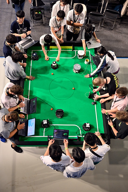 Teams gathered around a green RoboCupJunior soccer field, laptops open, small autonomous robots on the pitch.