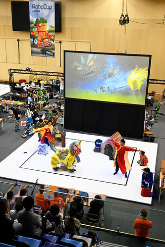 Costumed robots performing on a white RoboCupJunior OnStage floor in front of a projected backdrop and a seated audience.